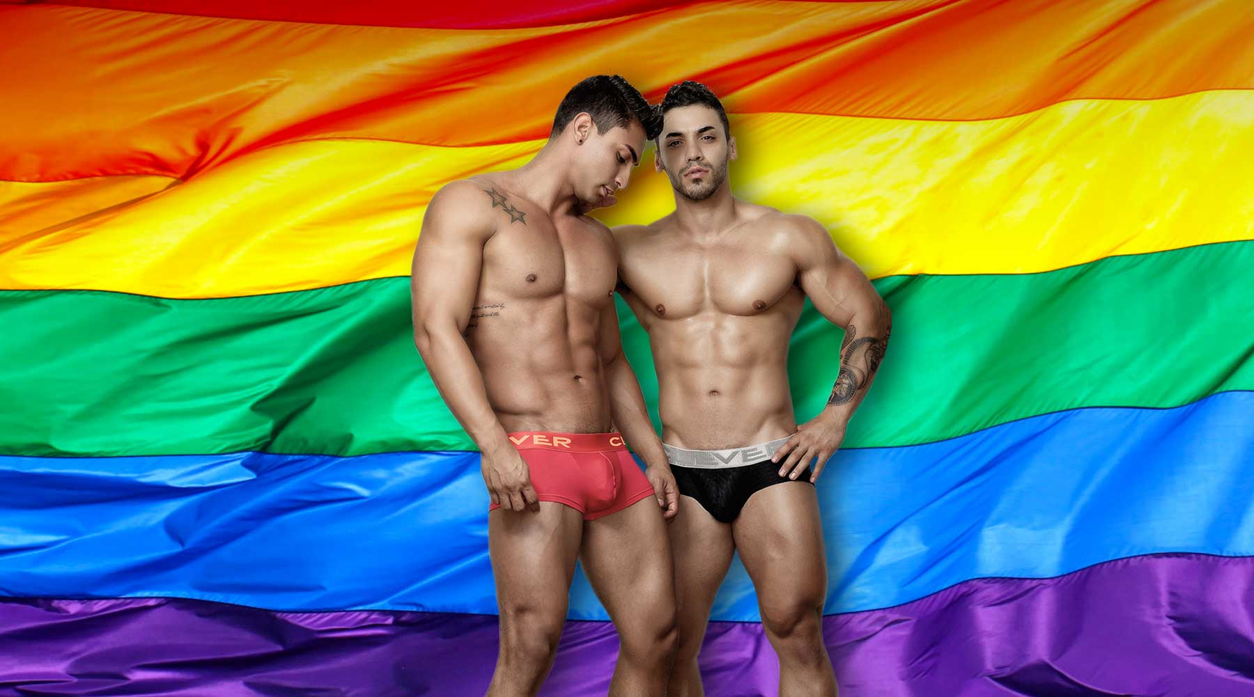 Two male underwear models standing in front of a Gay Pride flag.